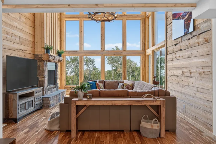 Great room featuring a two-story window wall with natural wood framing, a stone fireplace with a wood mantle, wood-finish flooring, and horizontal wood paneling