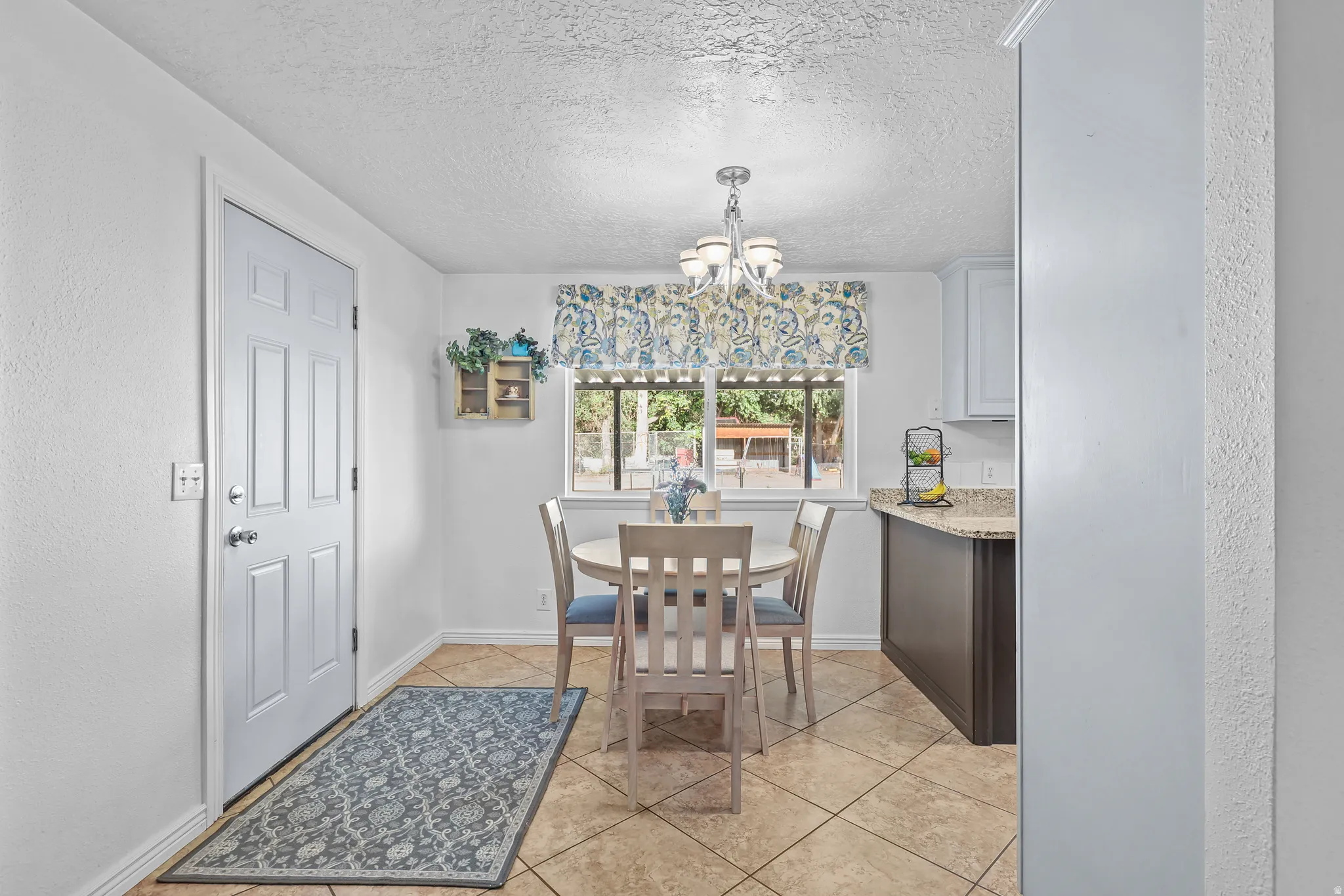 Tiled flooring throughout this dining area, featuring a window with an exterior view, a multi-light chandelier, a paneled door, and a partial view of kitchen cabinetry with a light-colored countertop