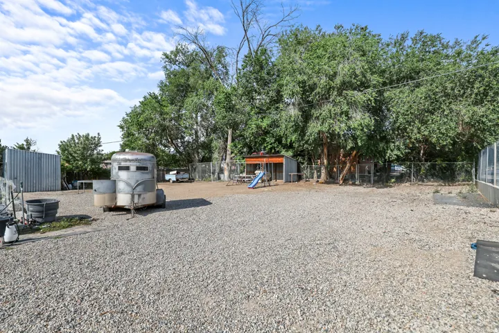 Expansive gravel yard featuring mature trees, a chain-link fence, and a utility shed