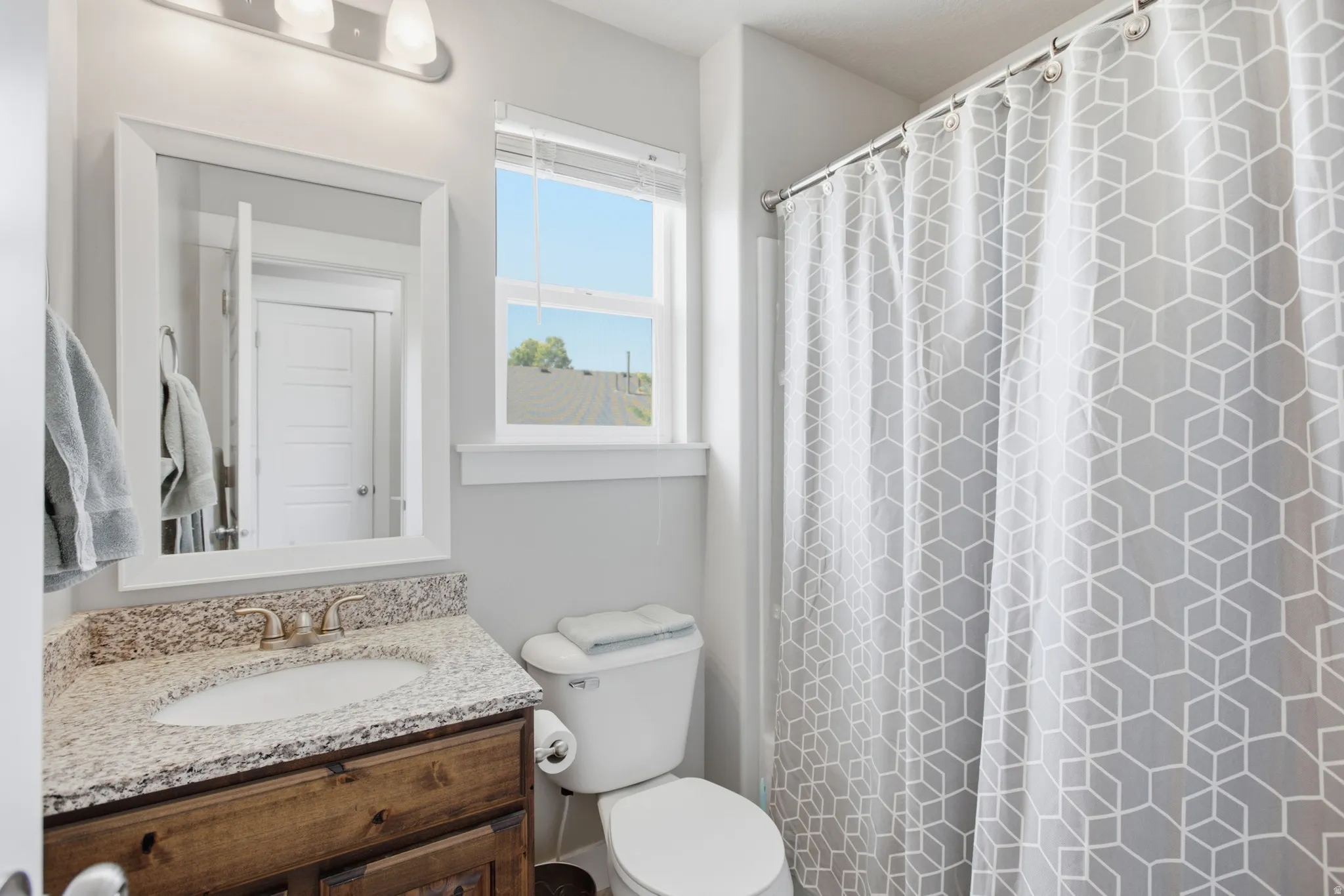 Full bathroom featuring a wood-finish vanity with a light-colored speckled countertop and undermount sink, a white-framed mirror, and a window with blinds