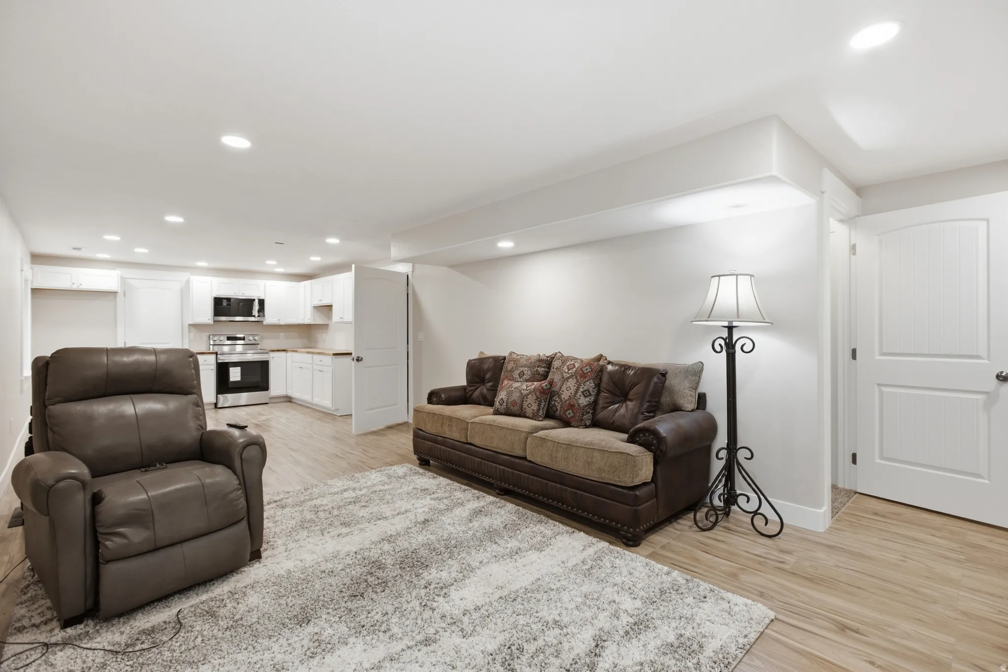 Open-concept living area with wood-finish flooring, recessed lighting, white paneled doors, and a full kitchen featuring white cabinetry, stainless steel appliances, and light countertops