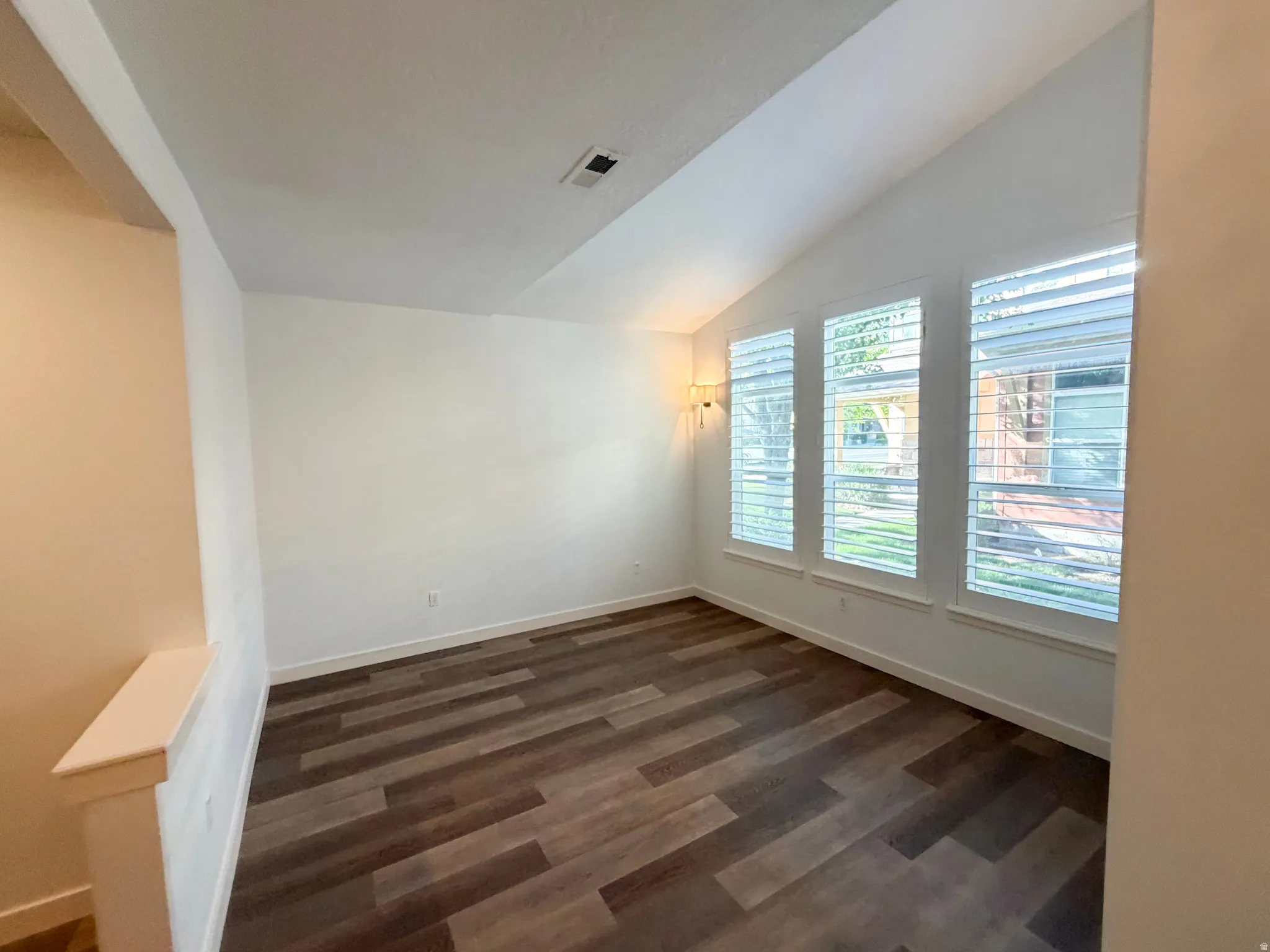 Vaulted ceiling room featuring three windows with plantation shutters, wood-finish flooring, and a wall-mounted sconce