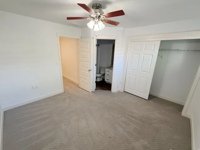Bedroom featuring light-toned carpeting, white walls, a ceiling fan with light fixture, sliding closet doors, and an entry to a bathroom with a vanity