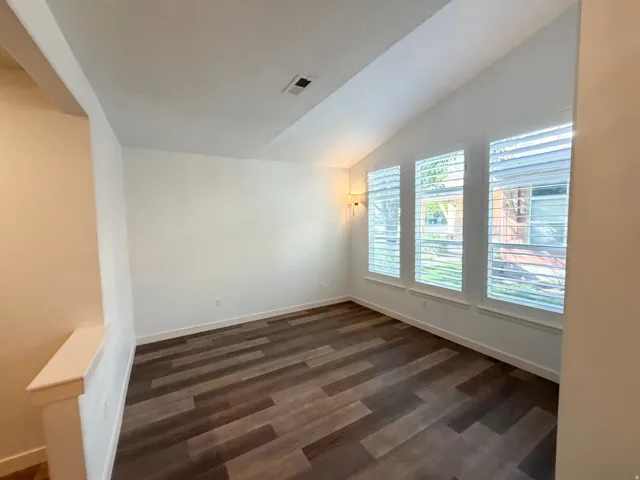 Vaulted ceiling room featuring three windows with plantation shutters, wood-finish flooring, and a wall-mounted sconce