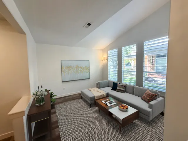 Vaulted ceiling living space featuring three tall windows with louvered blinds, wood-finish flooring, and a contemporary wall sconce