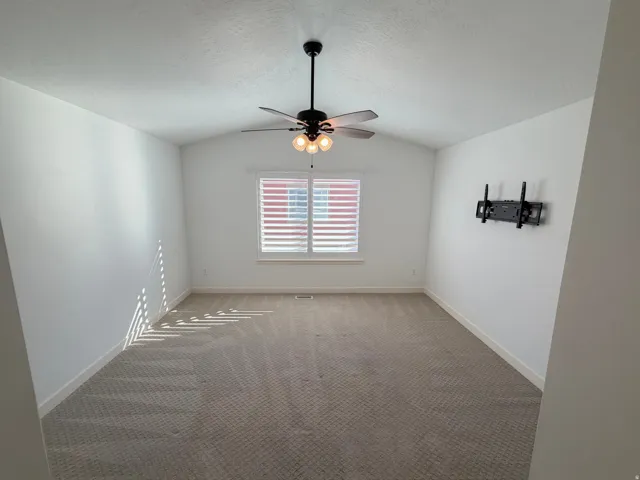 Spacious room featuring vaulted ceilings, a ceiling fan with light fixture, window with white plantation shutters, and plush patterned carpeting