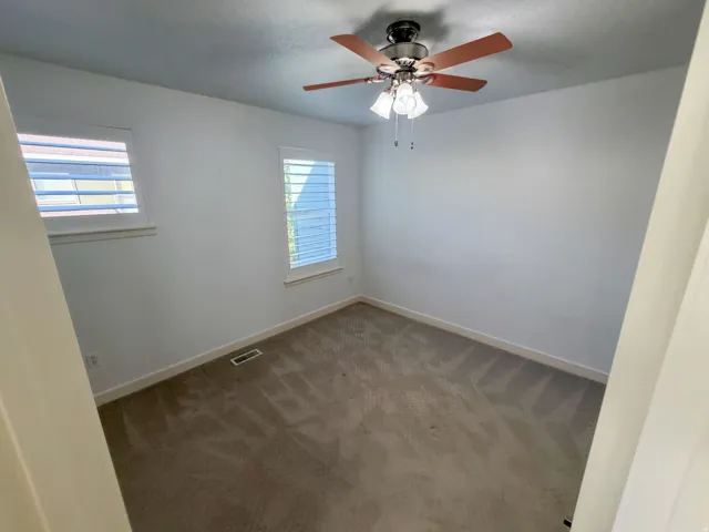 Room featuring a ceiling fan with integrated lighting, two windows with white plantation shutters, and light-toned carpet flooring