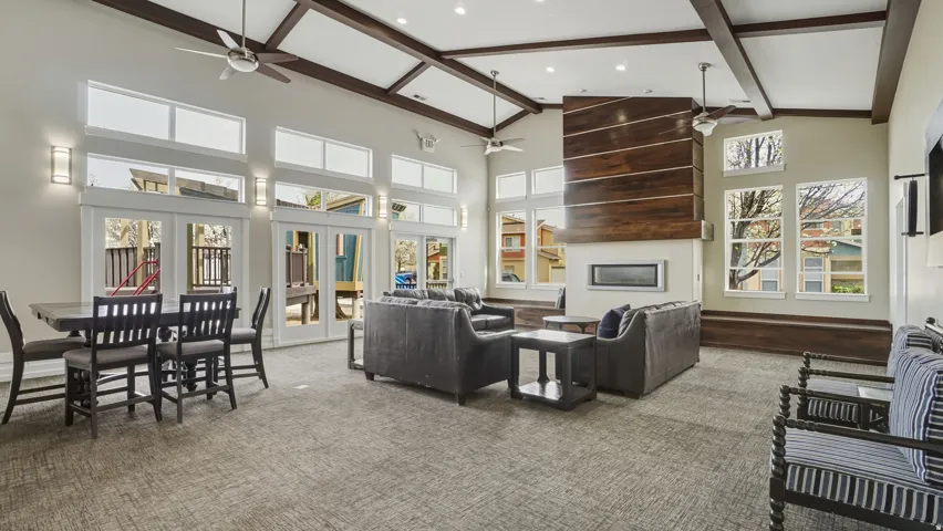 Vaulted ceiling with exposed beams, wood-paneled fireplace surround, multiple large windows, recessed linear fireplace, and ceiling fans