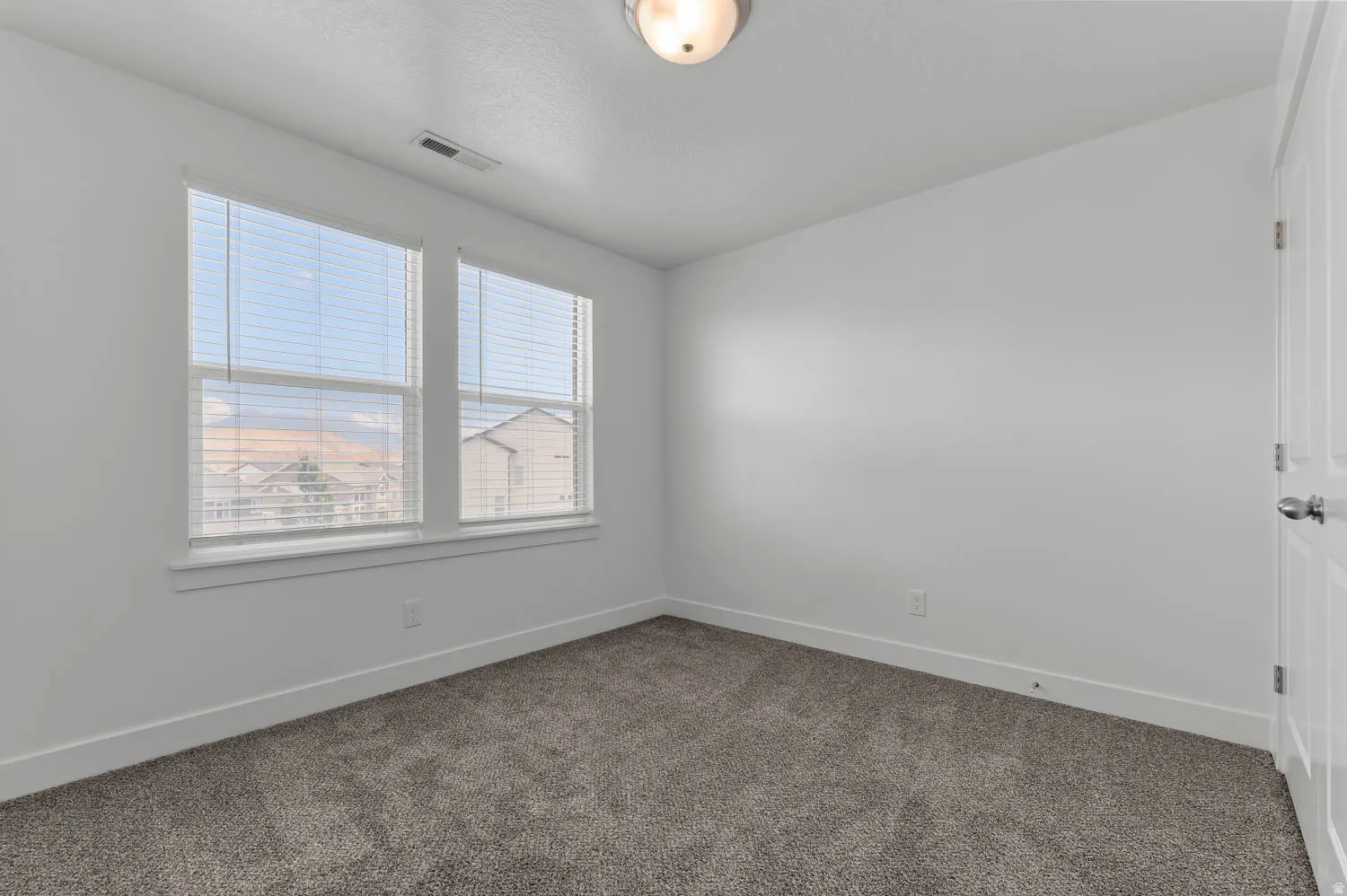 Light-filled interior featuring two windows with blinds, neutral wall paint, gray textured carpeting, and white baseboards