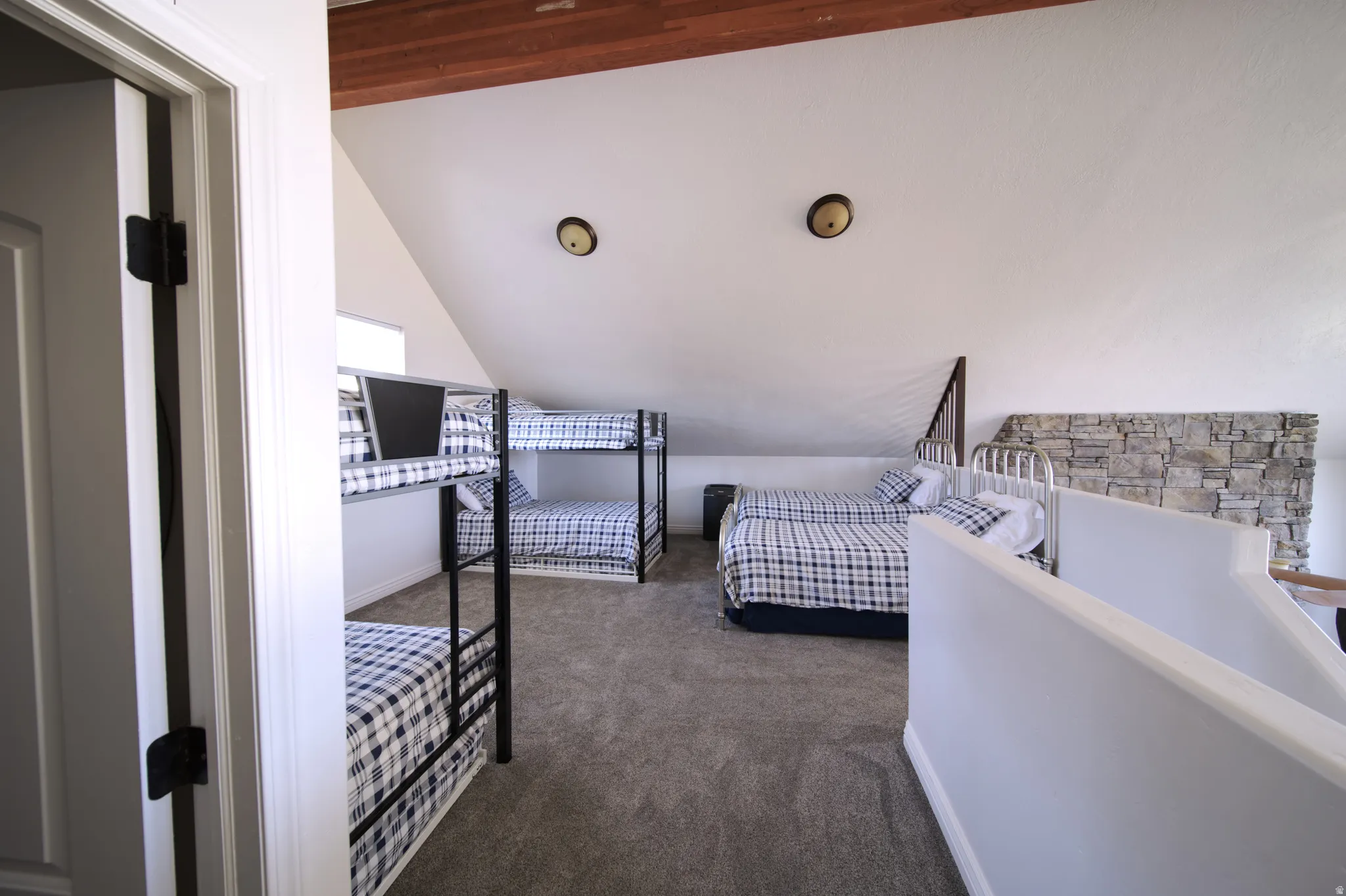 Carpeted loft space featuring an exposed wood beam ceiling, white walls, two wall-mounted light fixtures, and a stone accent wall