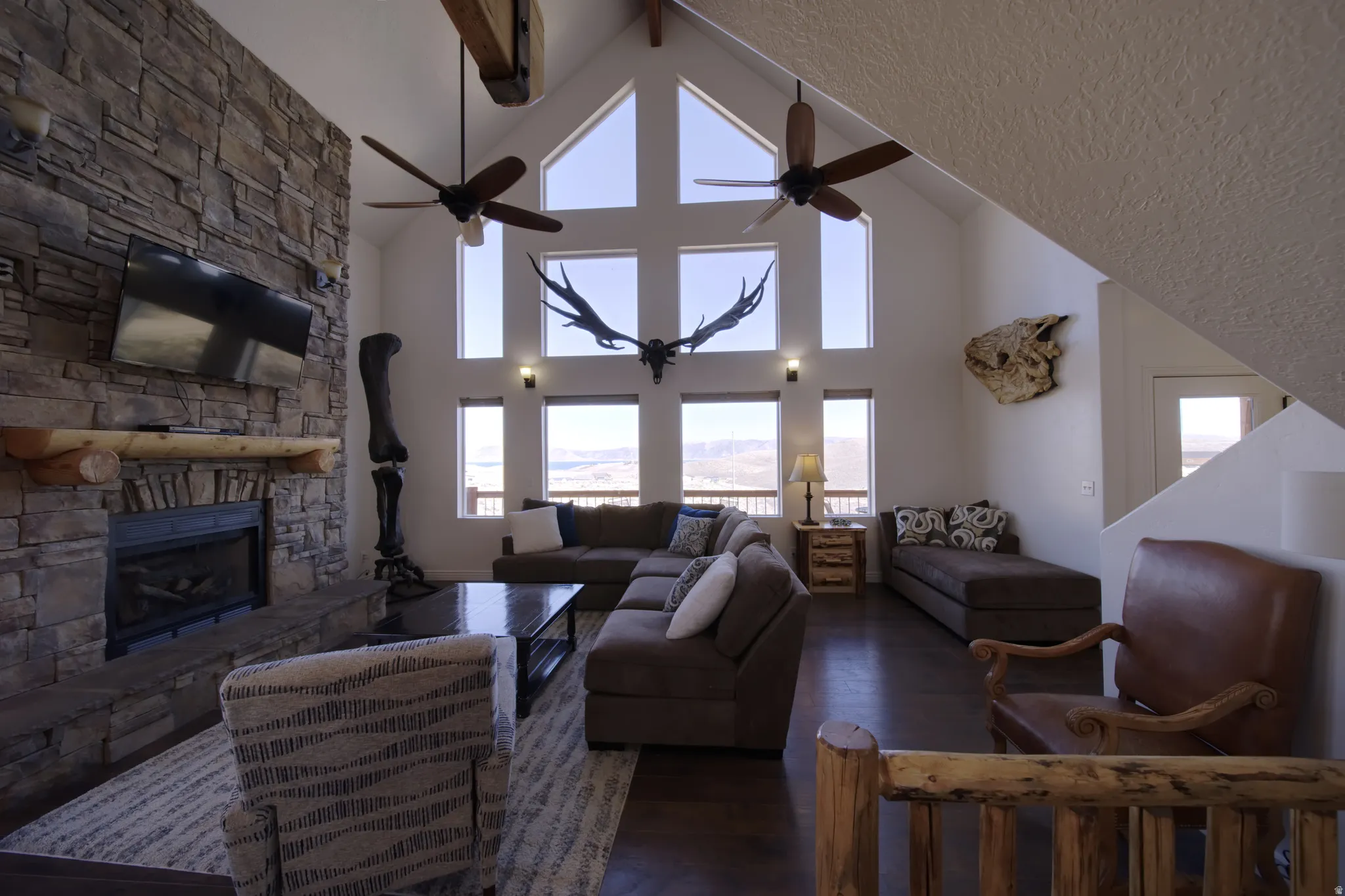 Spacious living area featuring a floor-to-ceiling stone fireplace with a rustic wood mantel, vaulted ceilings with exposed wood beams, and a window wall providing abundant natural light