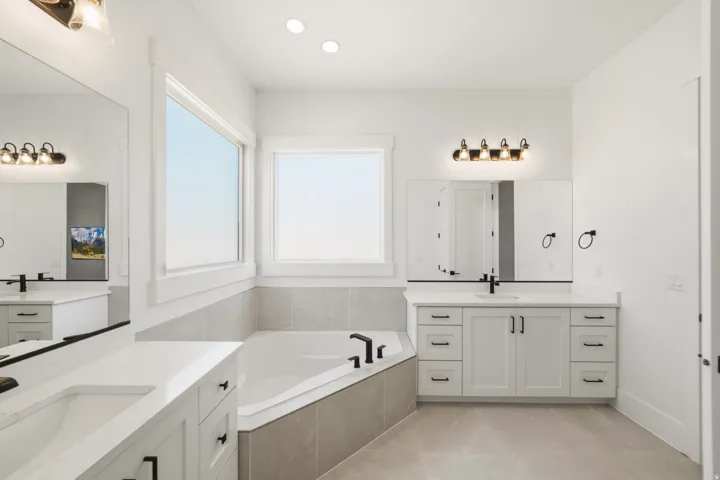Spacious bathroom featuring a built-in soaking tub with surrounding tile, dual vanity stations with integrated sinks and quartz countertops, and contemporary black fixtures