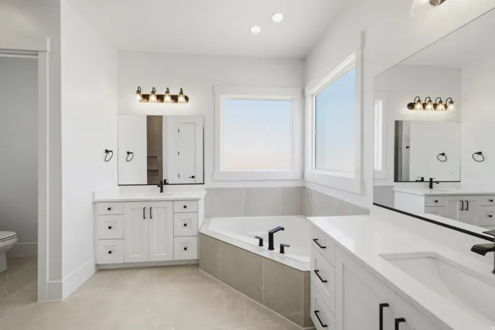 Spacious bathroom featuring a built-in corner soaking tub, dual vanity with white cabinetry and integrated sinks, contemporary black fixtures, and large windows