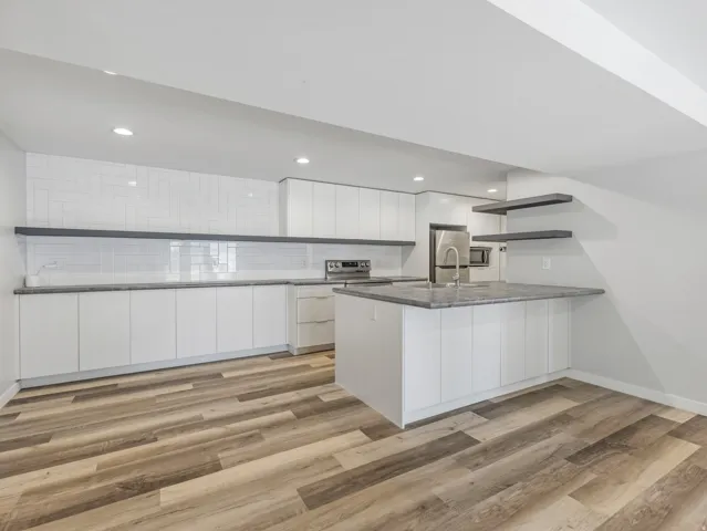 Modern kitchen featuring white flat-panel cabinetry, a central island with an undermount sink, stainless steel appliances, grey stone countertops, and wood-finish flooring