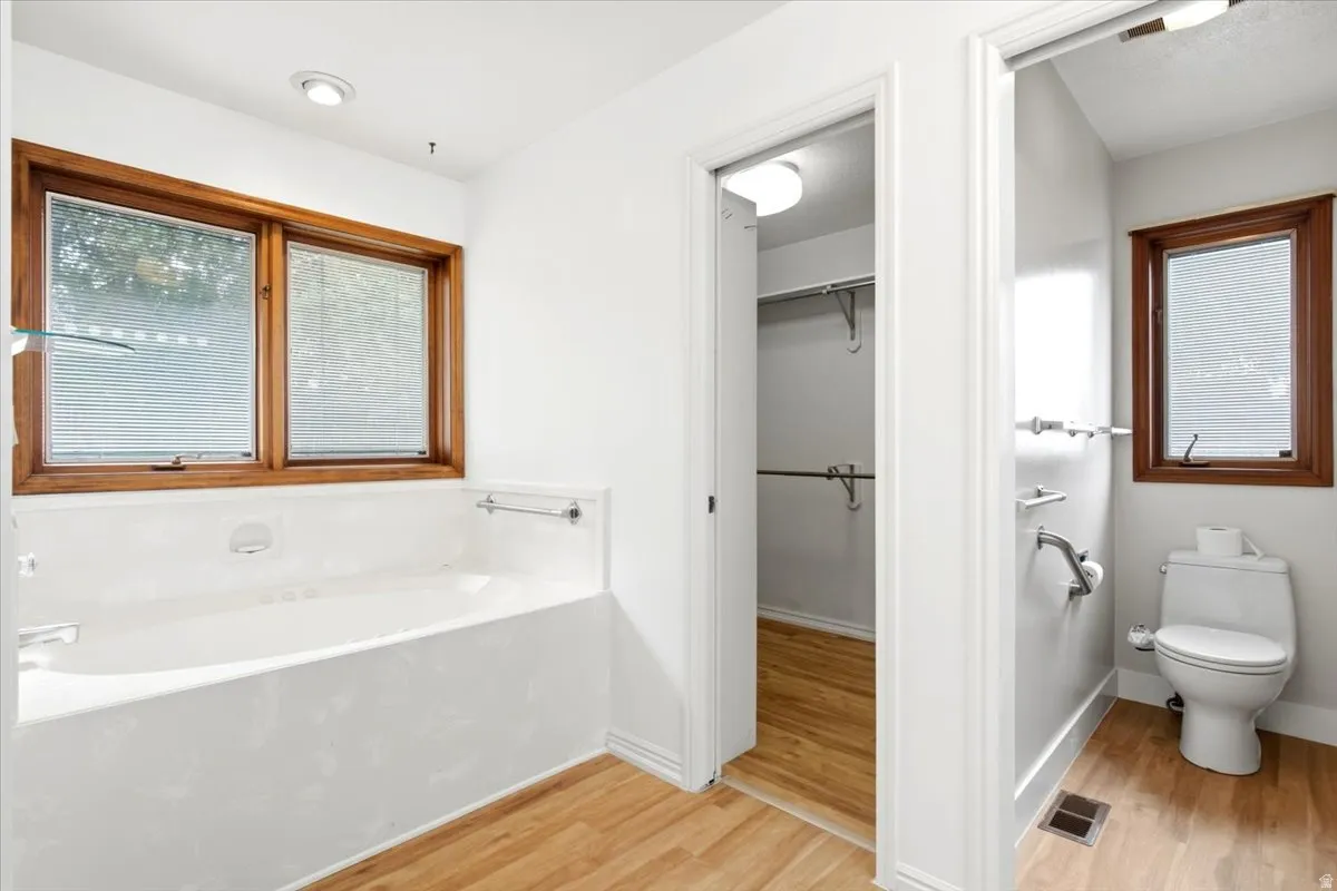 Bathroom featuring a built-in tub with dual wood-framed windows, wood-finish flooring, and a separate water closet with an additional window
