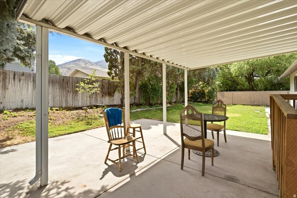 Covered patio with concrete flooring, supported by white posts, leading to a fenced backyard with mature trees