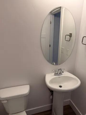 Pedestal sink with chrome fixtures and an oval mirror, complemented by wood-finish flooring and white baseboards