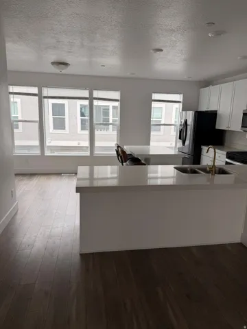Open-concept kitchen and living area featuring wood-finish flooring, a kitchen island with integrated sink, white cabinetry, a black refrigerator, and multiple windows with blinds