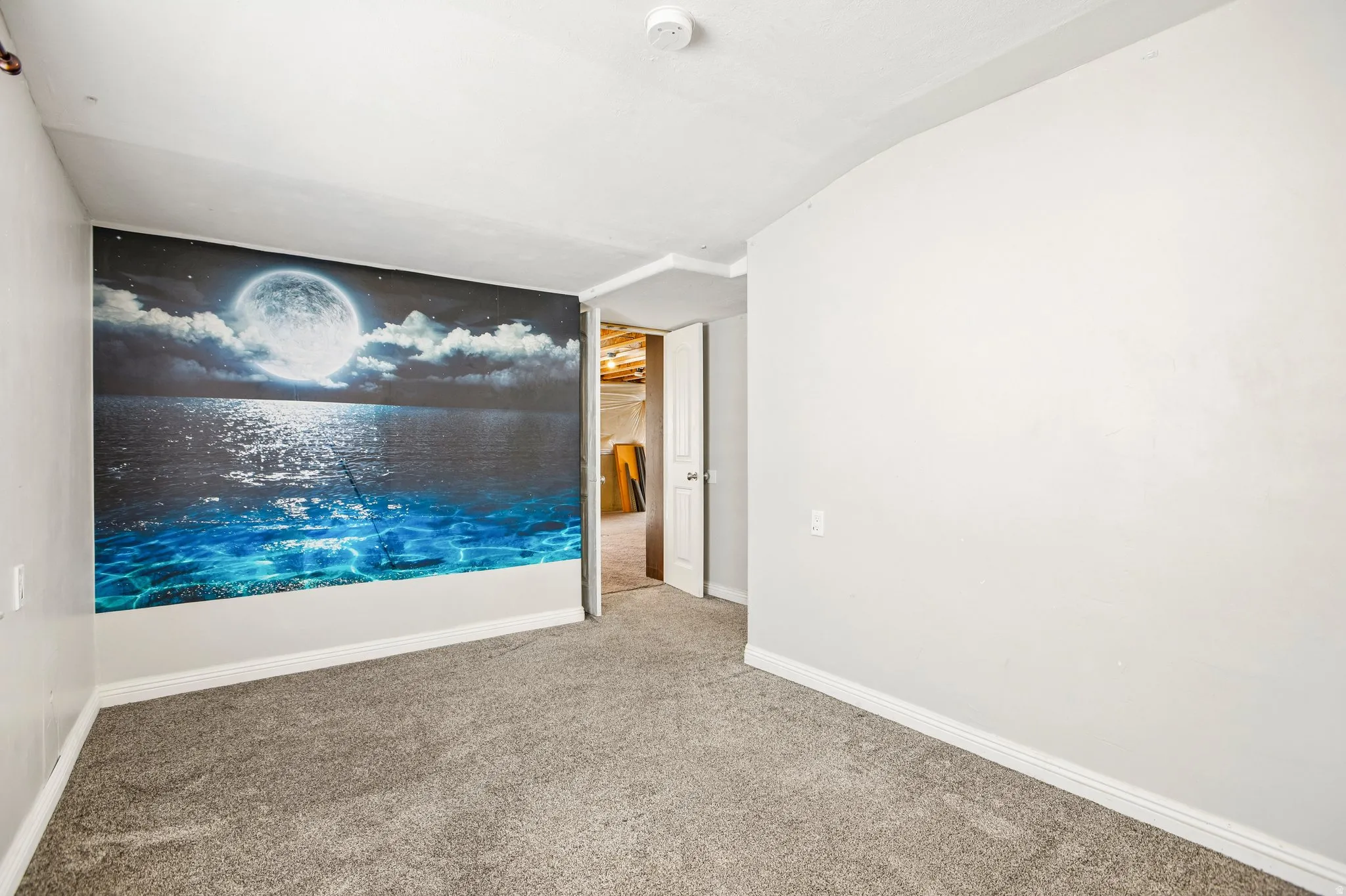 Spacious room featuring light gray walls, plush gray carpet, white baseboards, and a distinctive accent wall with a moonlit ocean mural