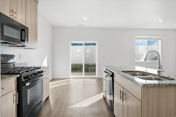 Open-concept kitchen featuring light wood-finish cabinetry, granite countertops, a stainless steel double basin sink with a high-arc faucet, and wood-finish flooring
