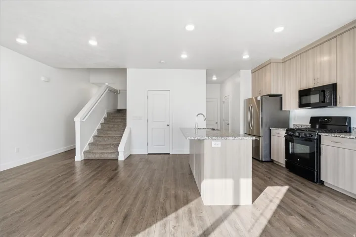 Open-concept living area featuring wood-finish flooring, a contemporary kitchen with light-toned cabinetry and granite countertops, stainless steel refrigerator, black range, and an island with an undermount sink