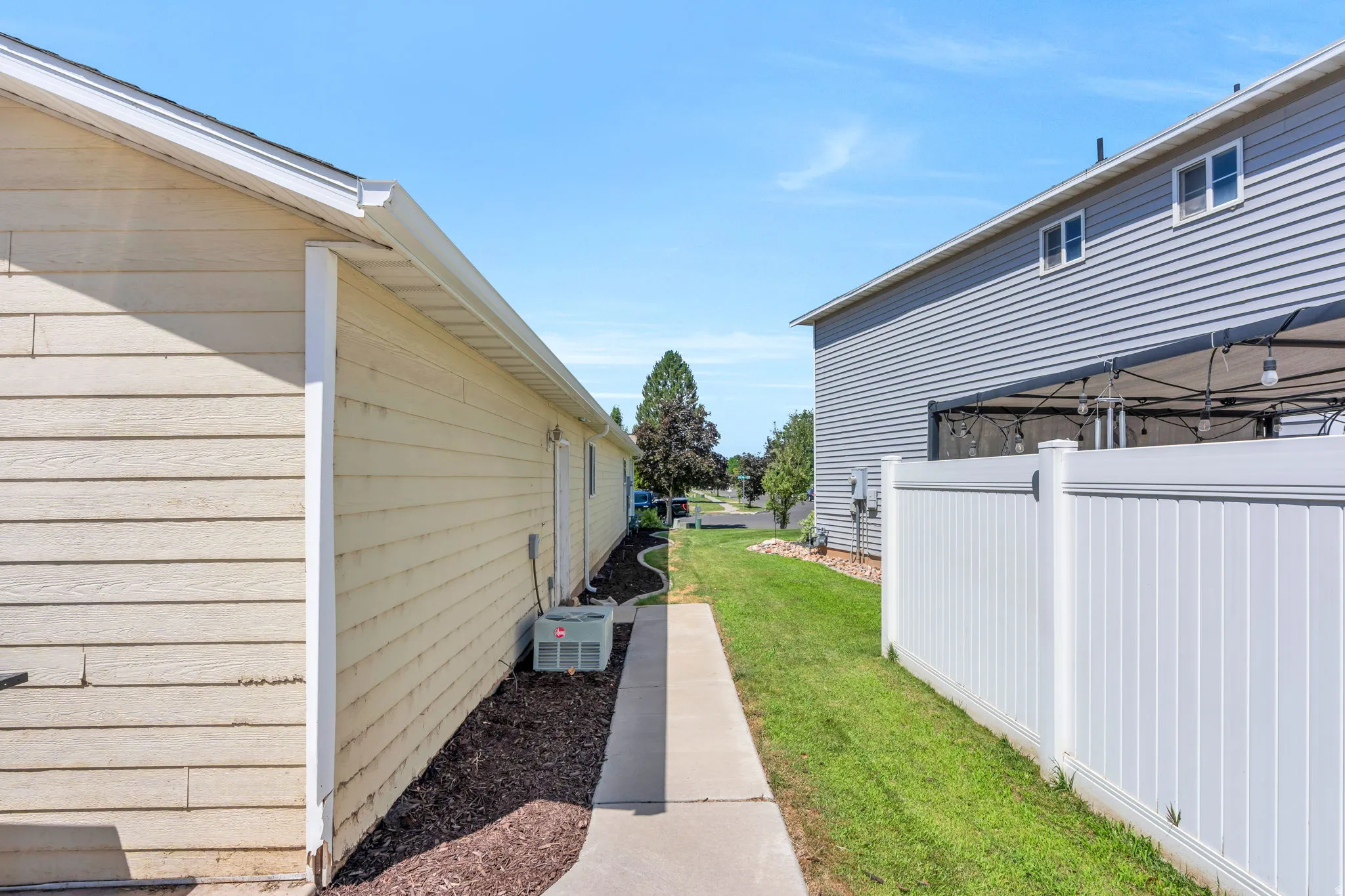 Exterior side passage featuring a concrete walkway, wood-finish siding, white vinyl fencing, and green lawn