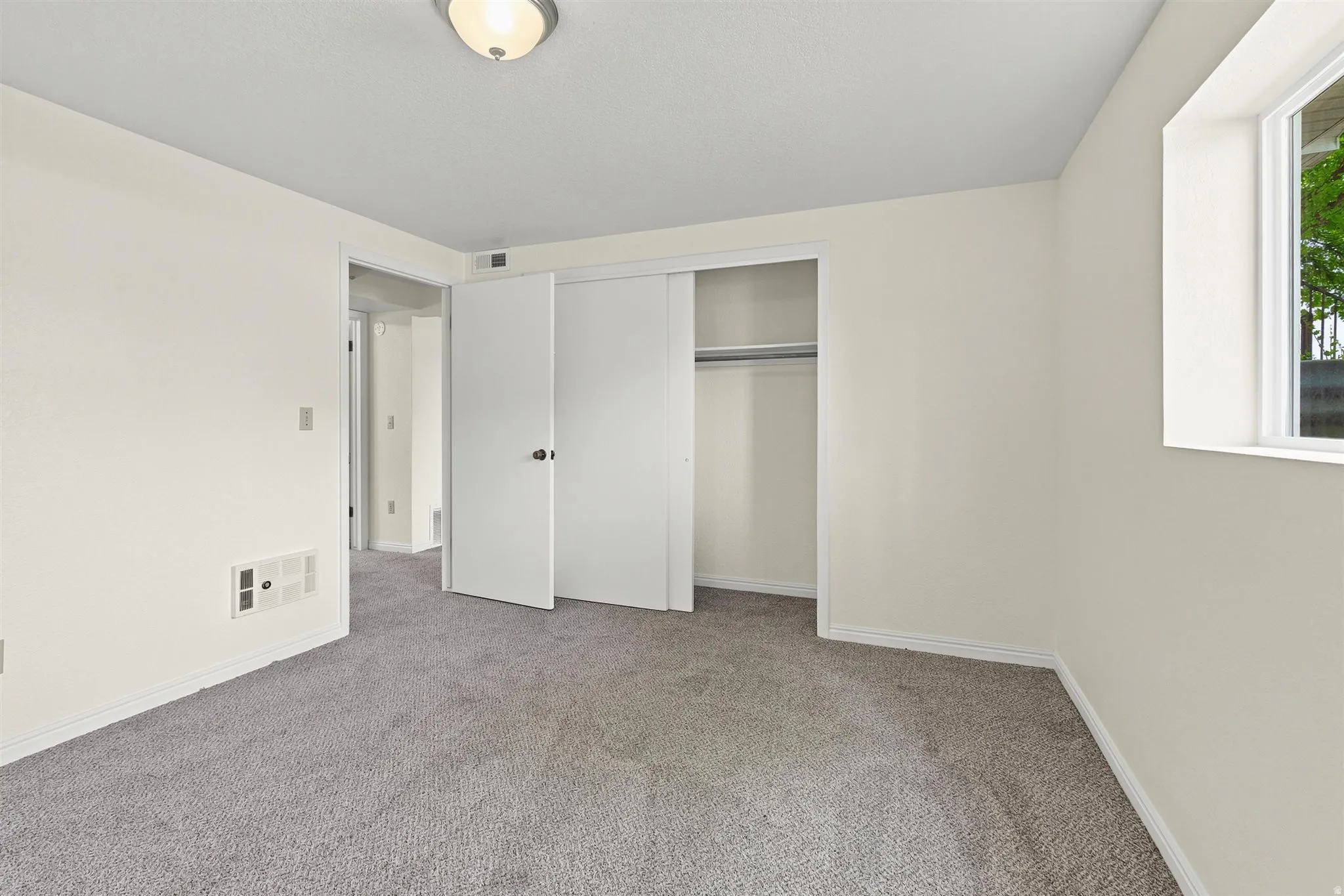 Spacious interior room featuring light gray carpet, white walls, white trim, a built-in closet with bi-fold doors, and a window providing natural light