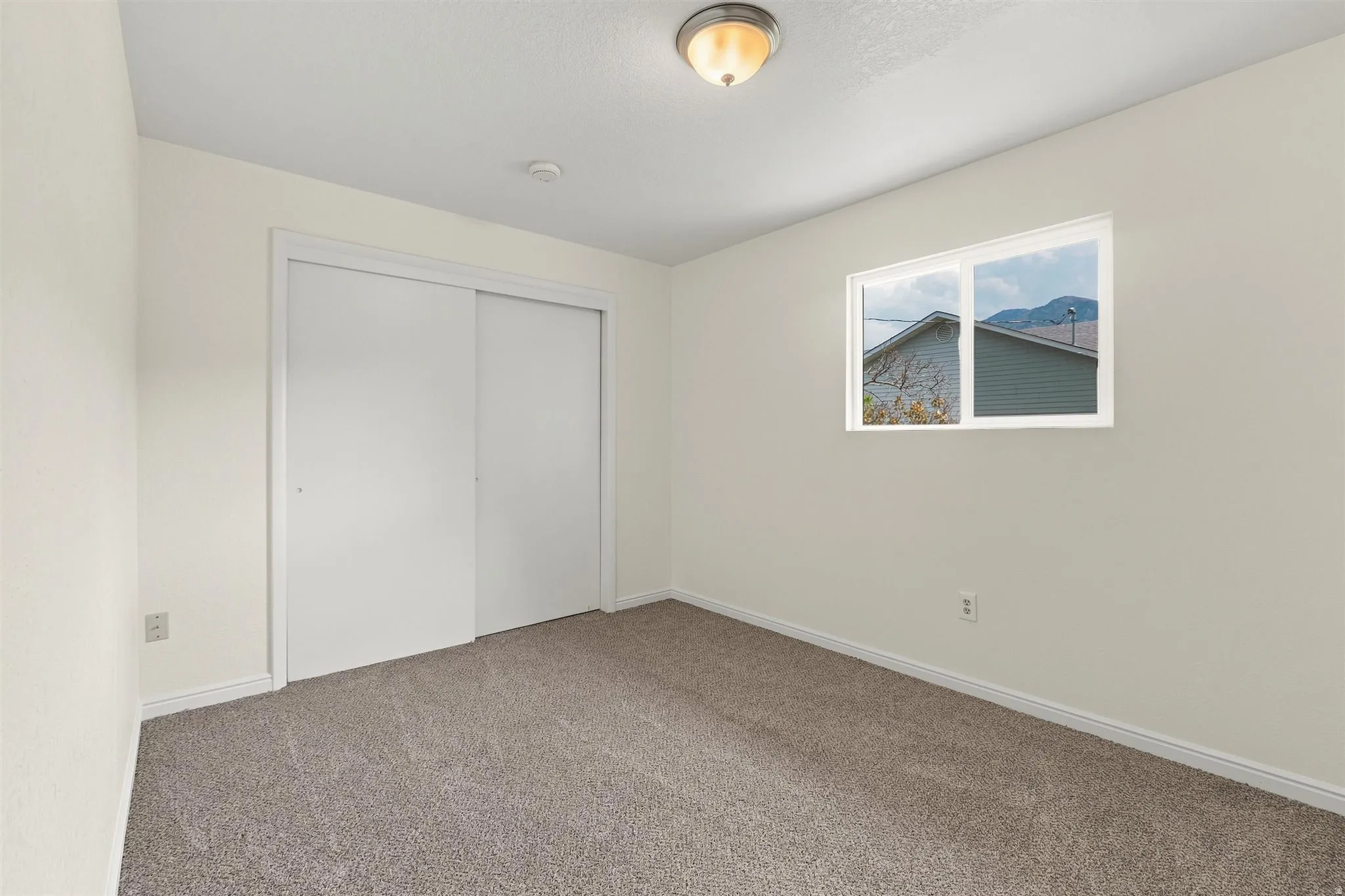 Neutral-toned bedroom featuring a dual-pane window, flush-mount ceiling light, built-in sliding door closet, and textured carpeting