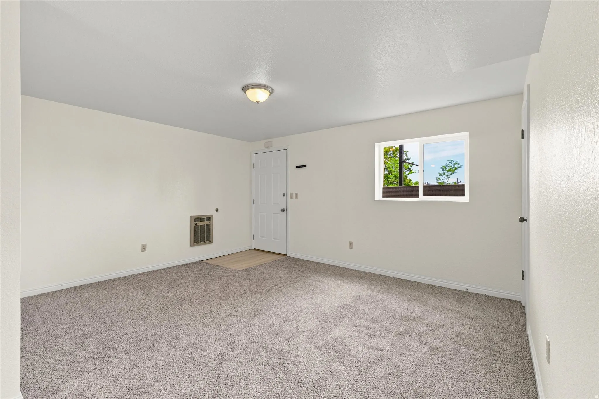 Carpeted room featuring a white door, a window with exterior visibility, a wall-mounted vent, a ceiling-mounted light fixture, and light-colored walls