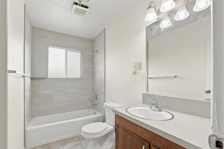 Bathroom featuring a tiled shower surround, frosted window, white bathtub, wood-finish vanity with an integrated sink, and overhead vanity lighting