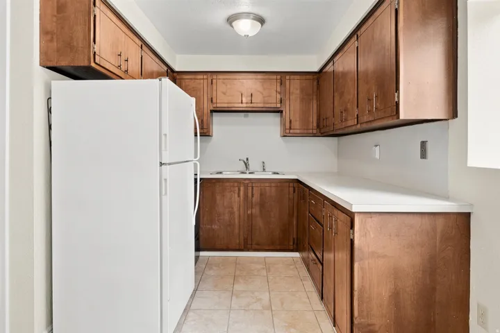 Compact kitchen featuring wood cabinetry, white countertops, a double basin sink, white refrigerator, and tile flooring