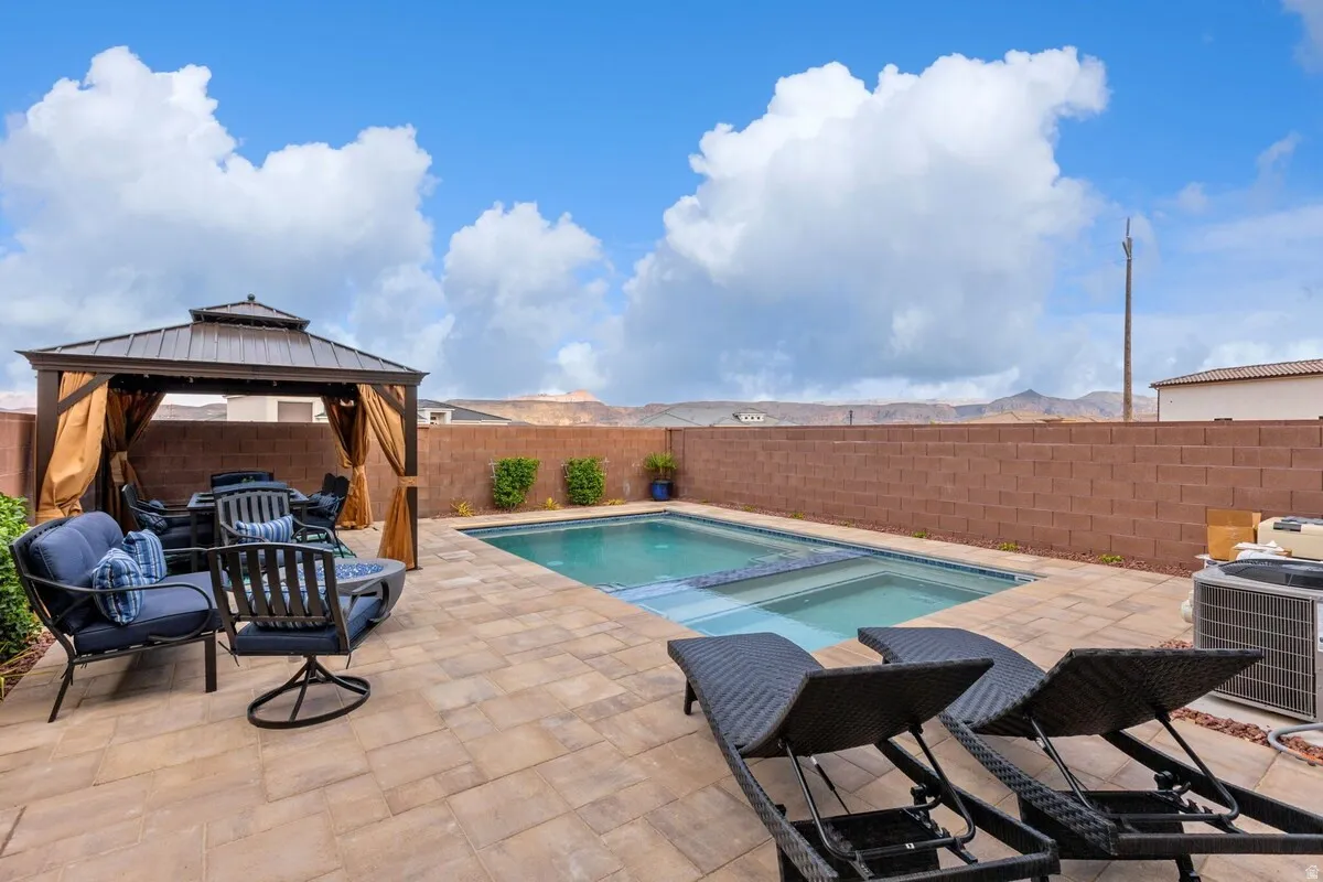 Backyard patio featuring a built-in swimming pool with an integrated spa, expansive paver decking, a metal-framed gazebo with fabric curtains, and a block perimeter wall
