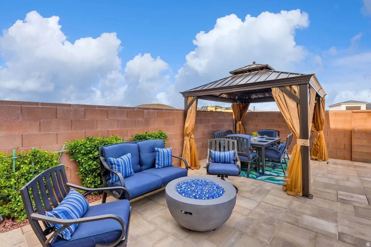 Expansive paver patio featuring a fire pit with blue glass accents, a dining gazebo with privacy drapes, and a low-maintenance block wall perimeter
