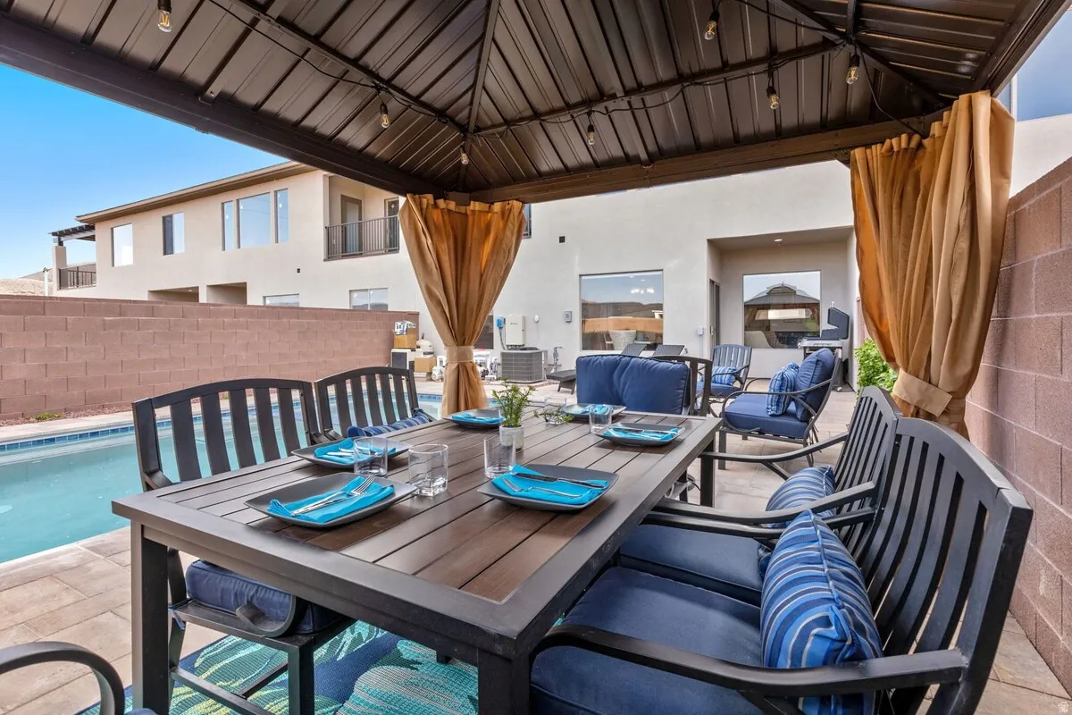 Covered patio with a metal gazebo roof and string lighting, featuring a dining table, seating, and a swimming pool