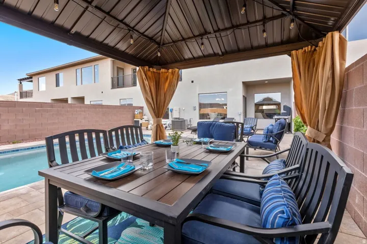 Covered patio with a metal gazebo roof and string lighting, featuring a dining table, seating, and a swimming pool