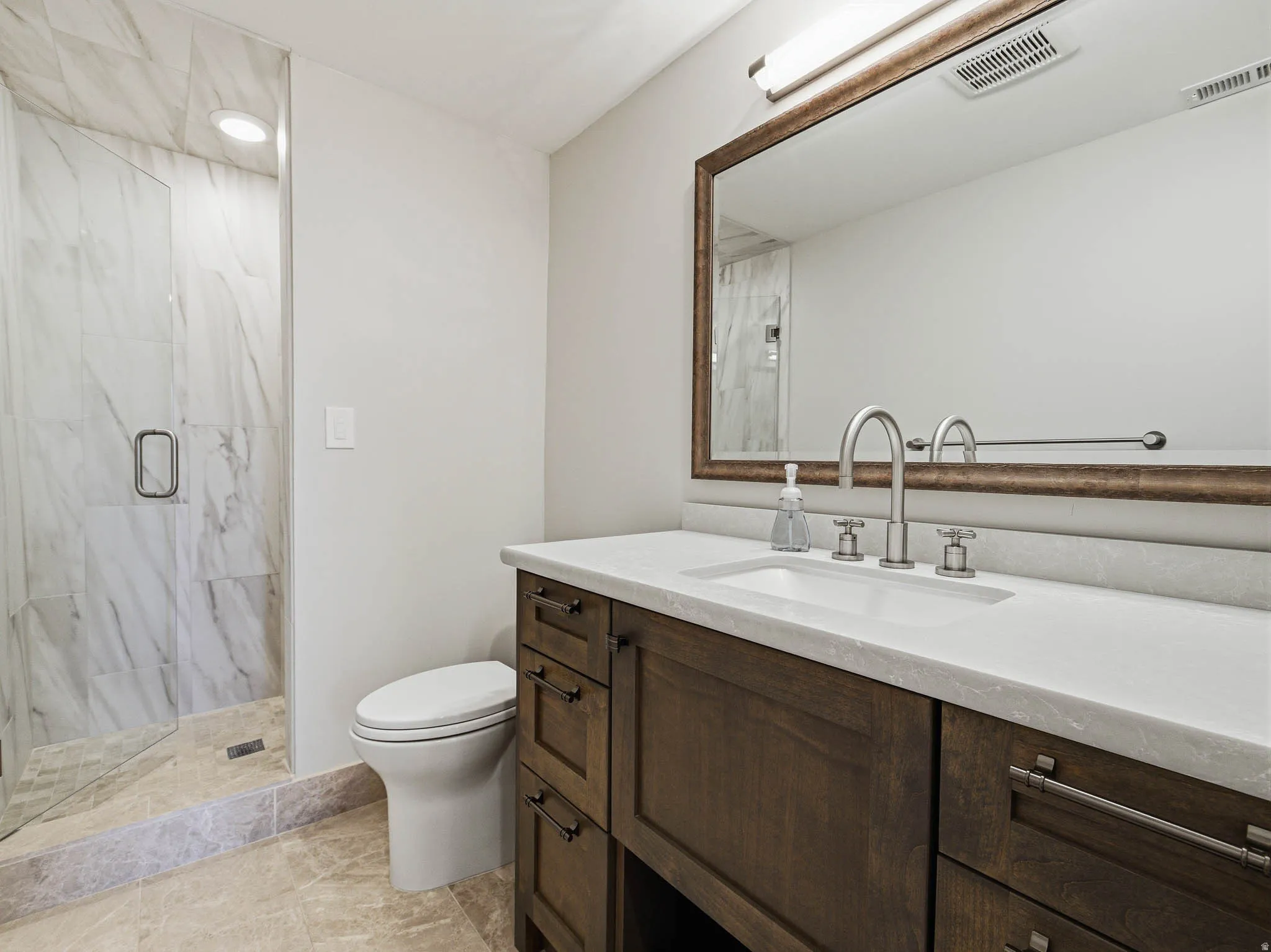 Full bathroom featuring a frameless glass shower enclosure with marble-look tile, a single vanity with a white countertop and dark wood cabinetry, and brushed nickel hardware
