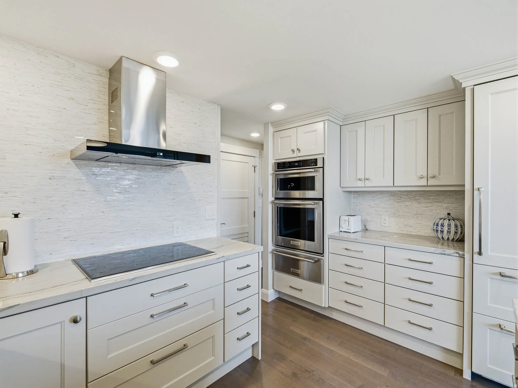 Contemporary kitchen featuring a glass cooktop, stainless steel range hood, double wall ovens, white shaker-style cabinetry, and light-toned countertops