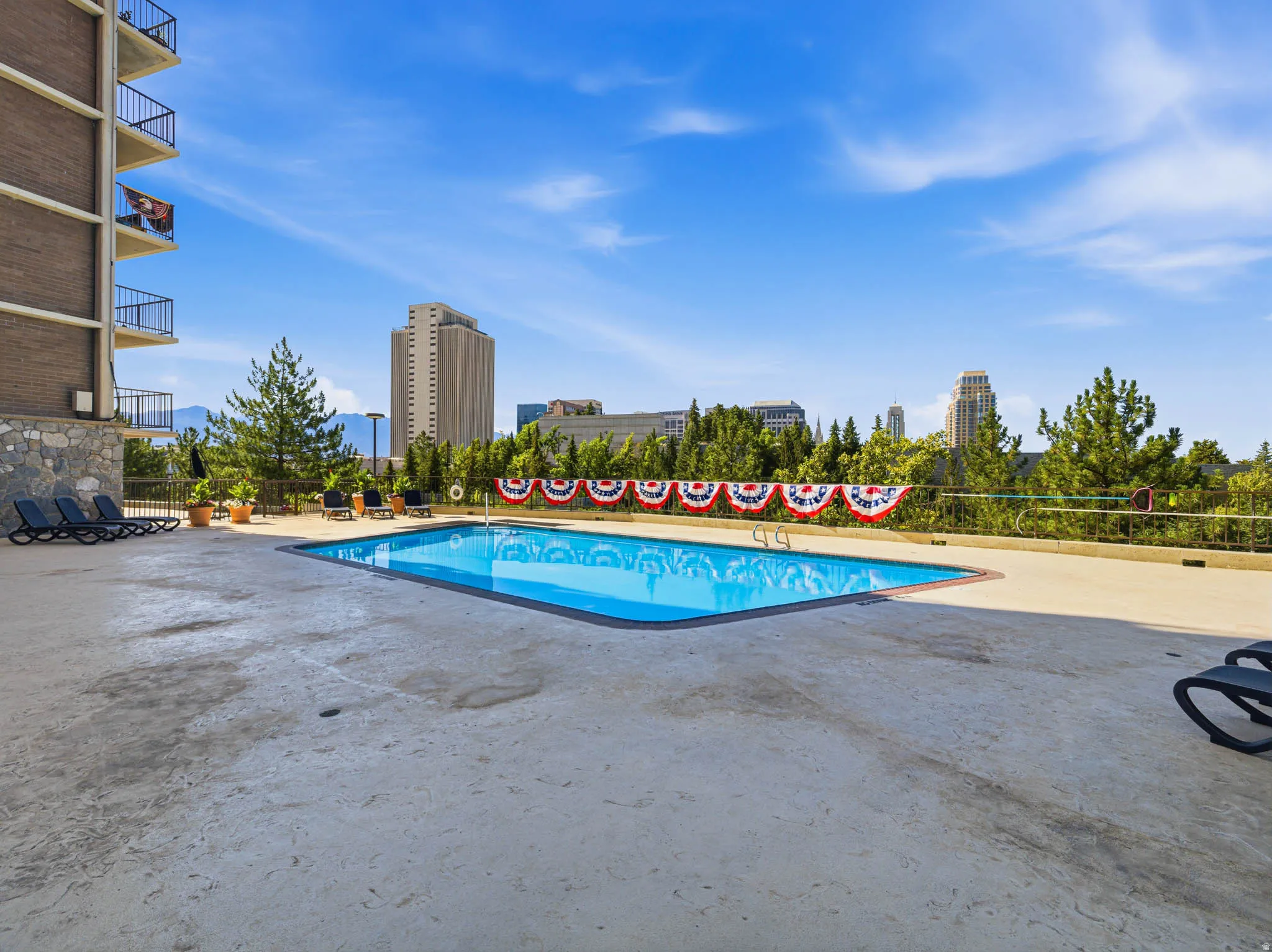 Outdoor pool with concrete deck, surrounded by mature trees, featuring a building exterior with balconies and distant city structures