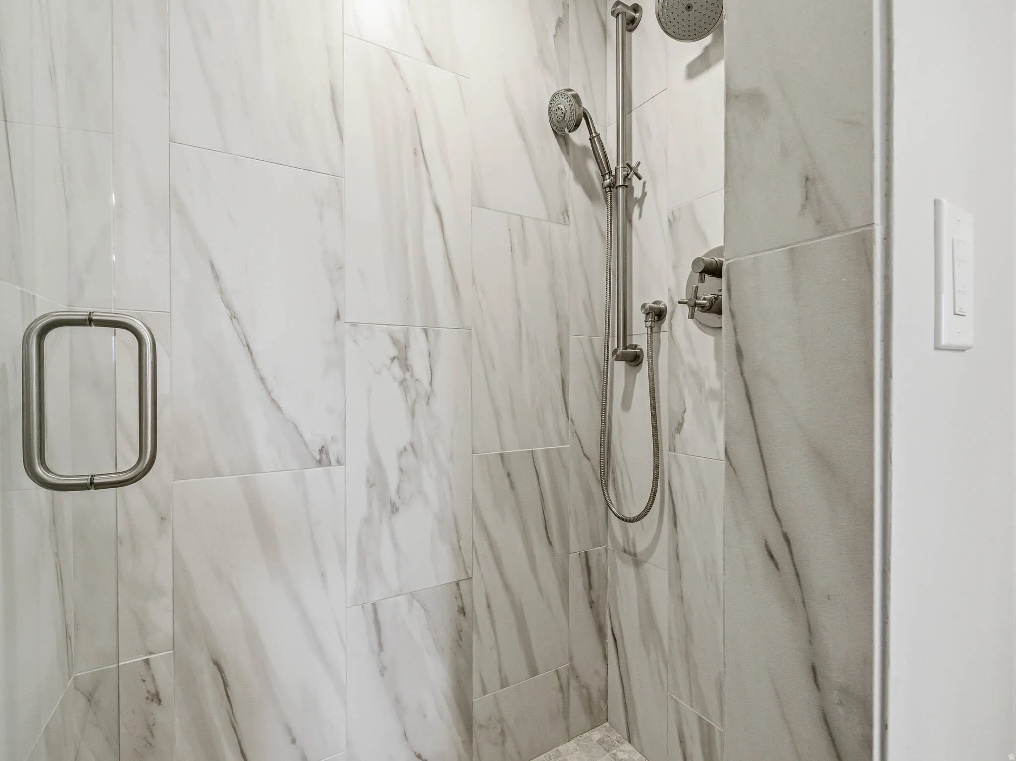 Spacious shower featuring floor-to-ceiling marble-look tile, a frameless glass door with brushed nickel hardware, and a dual shower head system