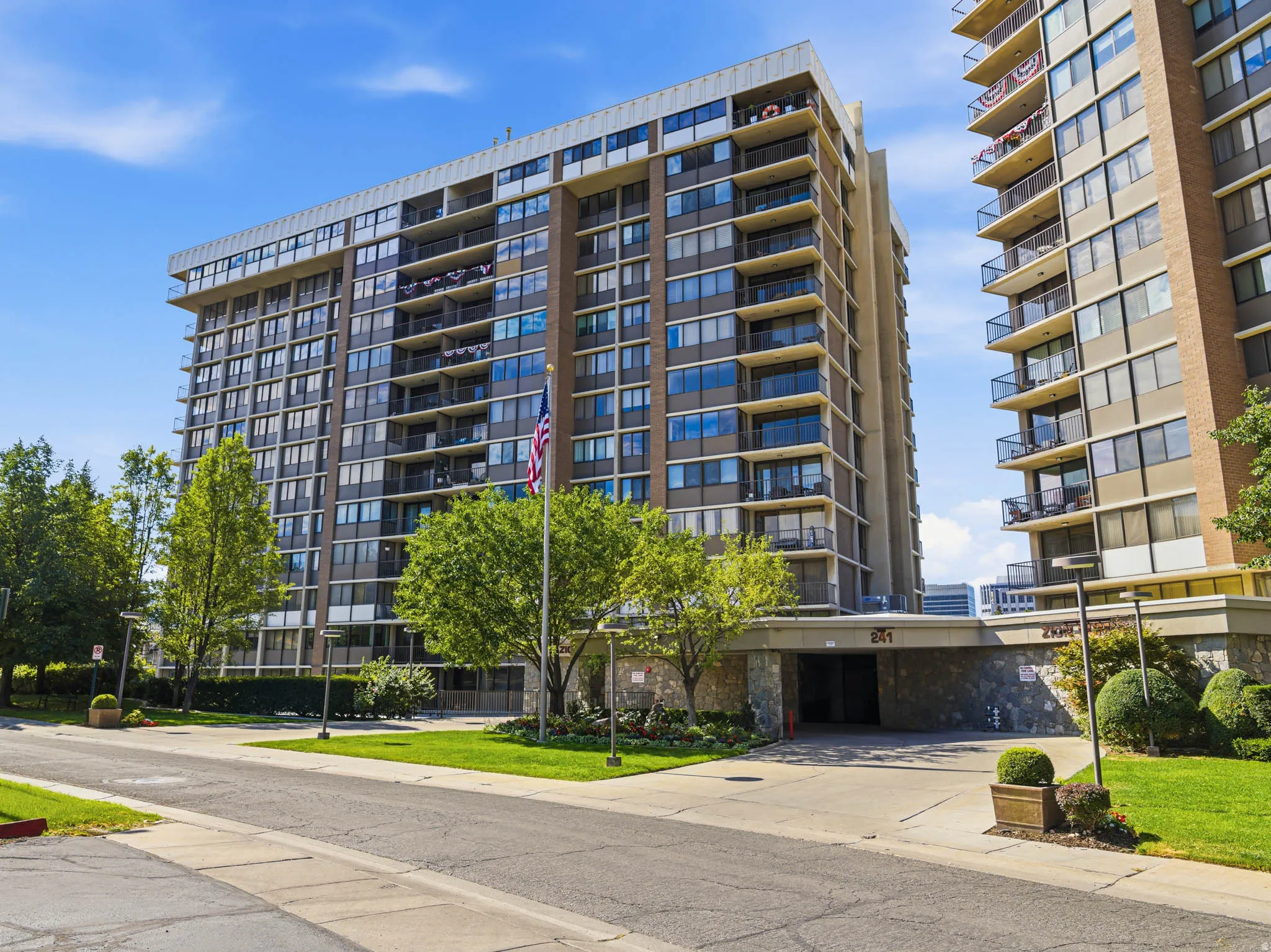 Multi-story building featuring numerous private balconies with black railings, a stone-clad entrance, and a well-maintained grassy frontage