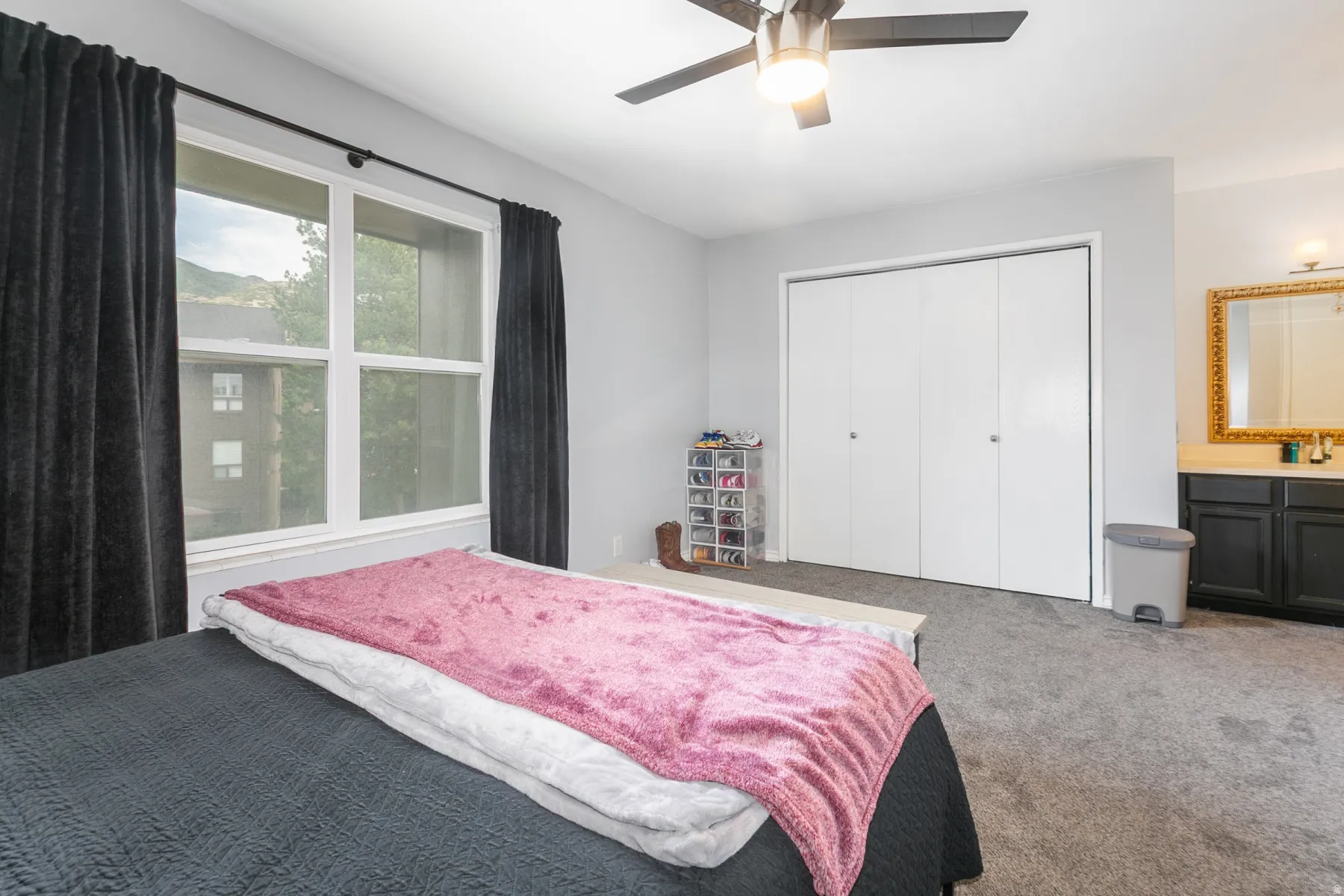 Bright bedroom featuring a large window, ceiling fan with integrated lighting, bi-fold closet doors, and built-in vanity with a gold-framed mirror