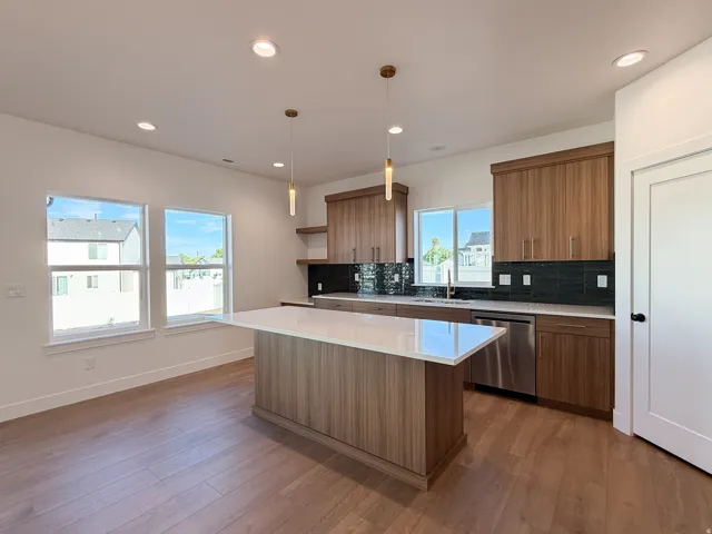 Spacious kitchen featuring an expansive island with a light-toned countertop, wood-finish cabinetry, dark tiled backsplash, and stainless steel appliances
