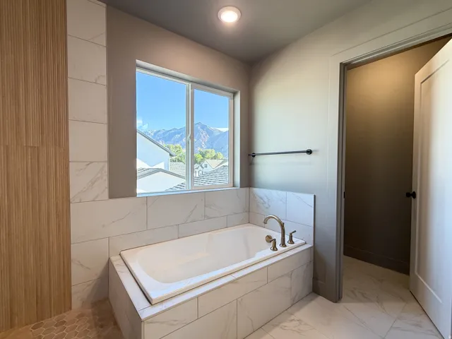 Built-in soaking tub with brass fixtures, marble-look tile surround, and large window