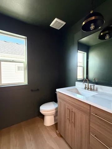 Wood-finish flooring, single vanity with a white countertop and integrated sink, modern pendant lighting, large wall mirror, and a window providing natural light