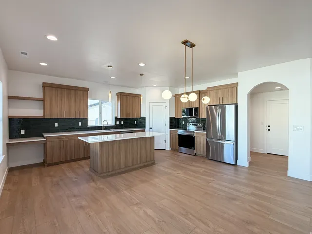 Open-concept kitchen and dining area featuring wood-finish cabinetry, stainless steel appliances, a large center island with white countertop, black tile backsplash, pendant lighting, and wood-finish flooring throughout the main living space