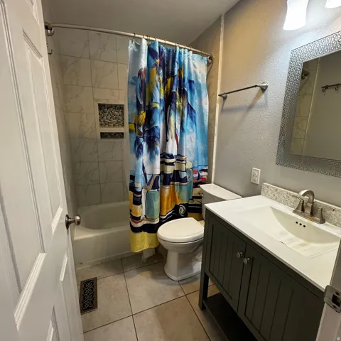 Bathroom featuring a white bathtub, tile shower with built-in shelf decorated with mosaic accent tiles, dark vanity with integrated sink, and granite backsplash, and a framed mirror