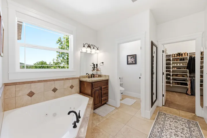 Soaking tub with tiled surround, dual-sink vanity with wood cabinetry and stone countertop, and separate water closet