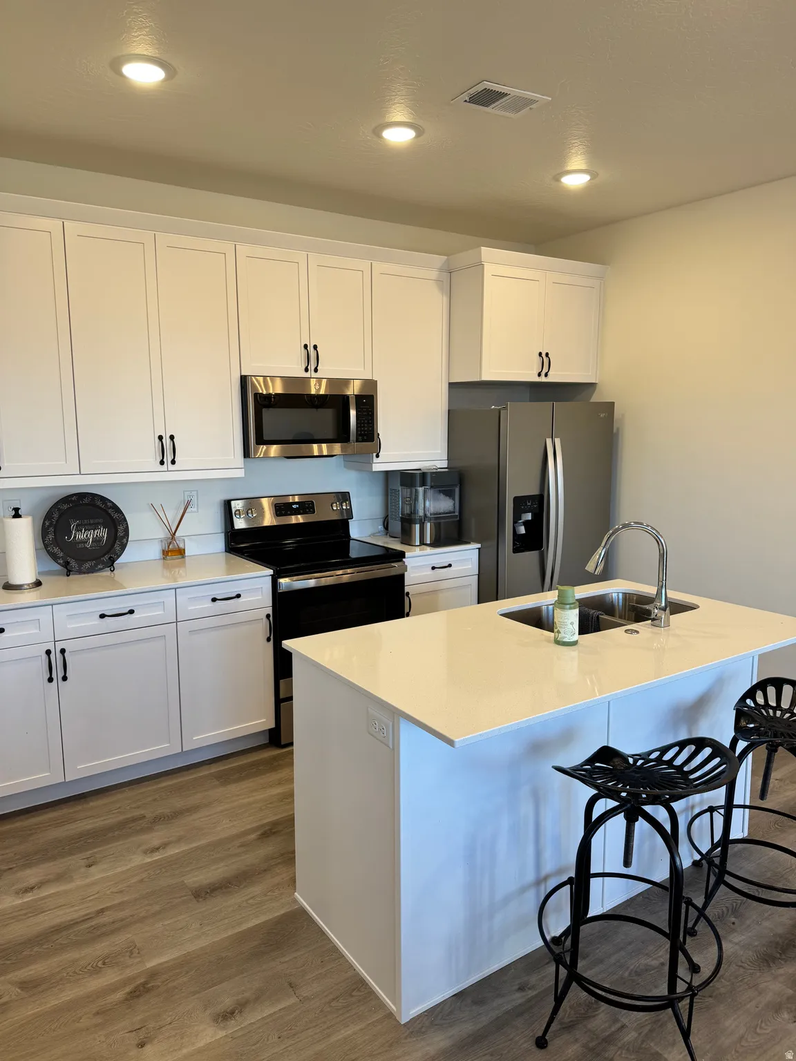 Contemporary kitchen featuring white cabinetry, stainless steel appliances, light-toned countertops, a central island with an undermount double sink, and wood-finish flooring