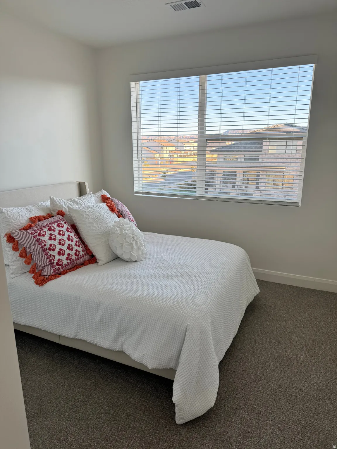 Carpeted room featuring a large window with horizontal blinds, offering distant views of residential rooftops