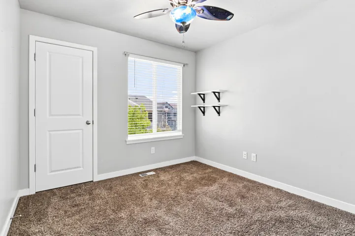 Carpeted room featuring a white paneled door, a window with blinds, two wall-mounted shelves, and a ceiling fan with integrated lighting